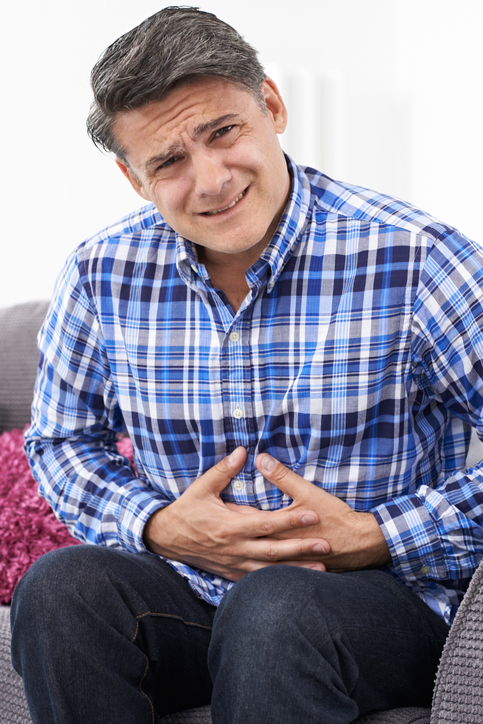 SII - hombre con dolor abdominal, calambres, sujetándose el abdomen, camisa azul a cuadros, sentado en el sofá