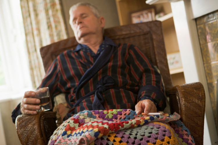 un abuelo dormido en una silla tapado con una manta, un vaso de alcohol y un cigarrillo en la mano