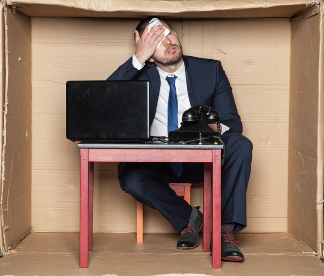 Hombre de negocios sentado en un escritorio dentro de una caja, espacio reducido, estrés laboral y aumento de la carga de trabajo, ordenador y teléfono, hombre secándose la frente.