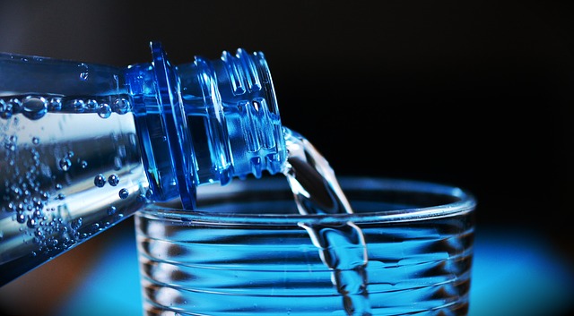 Se vierte agua de la botella al vaso. Sólo se ven el cuello de la botella y la parte superior del vaso. El fondo es oscuro y azul.