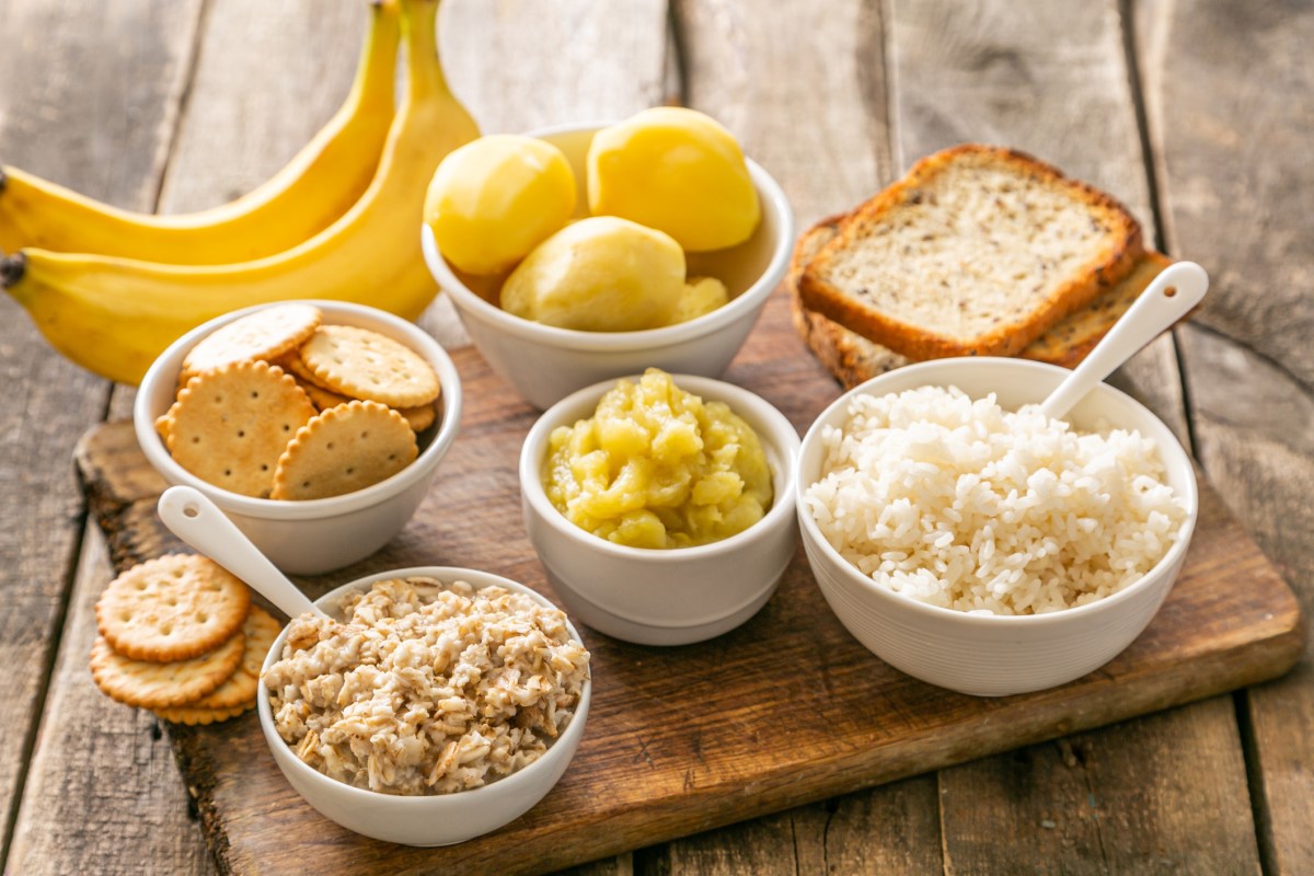 Ejemplo de una dieta adecuada para la diarrea aguda: arroz, patatas, plátanos, galletas, galletas saladas, en cuencos y sobre una tabla de cortar de madera.