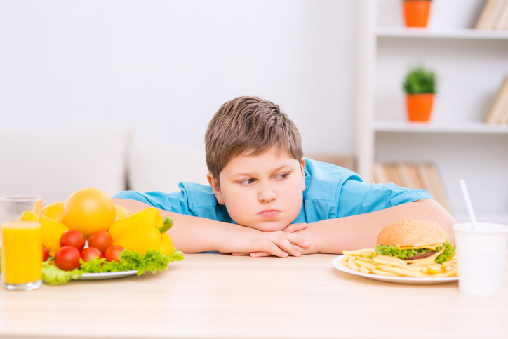 Un niño gordo entre dos platos de comida sana y poco sana.