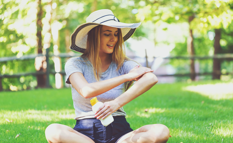 Una joven se pone crema solar, sentada en un prado, con un sombrero para protegerse la cabeza