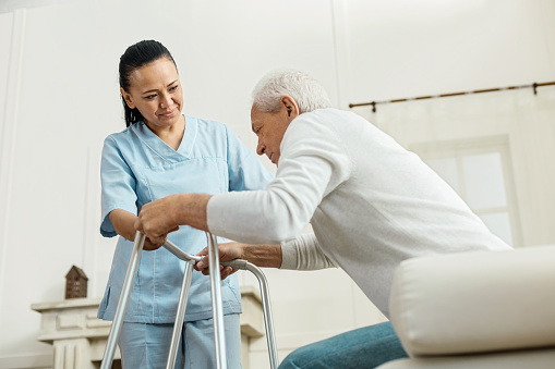 un anciano sentado en una cama de hospital intentando levantarse con la ayuda de un andador y una enfermera