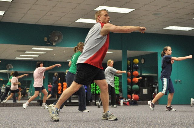 Hay cinco personas haciendo ejercicio en el gimnasio. Un hombre y cuatro mujeres. Van vestidos de forma atlética, de espaldas a un gran espejo. La pared es verde y la moqueta del suelo gris.