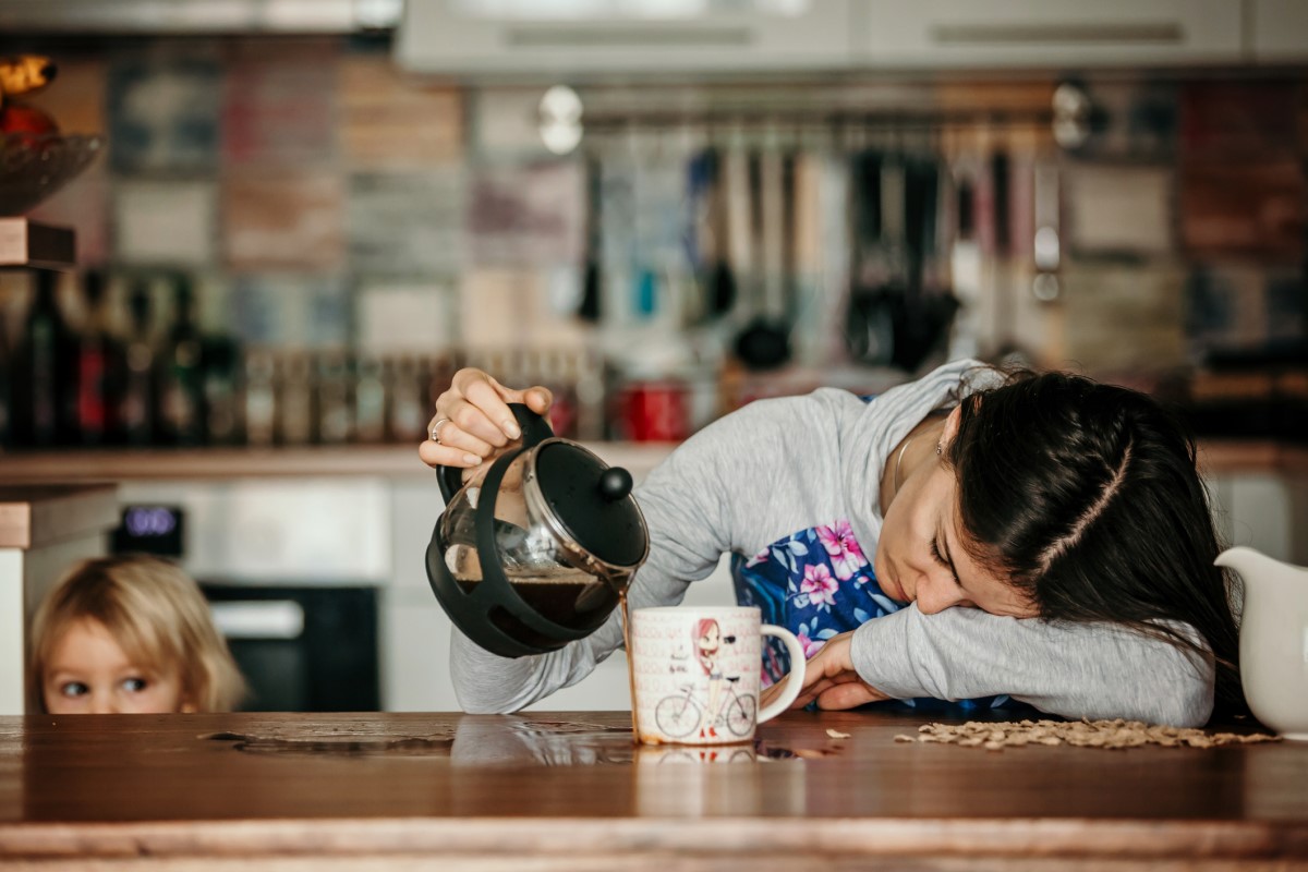 Una mujer cansada está tumbada en la encimera de la cocina, derramando café junto a una taza, un niño de pie a su lado.