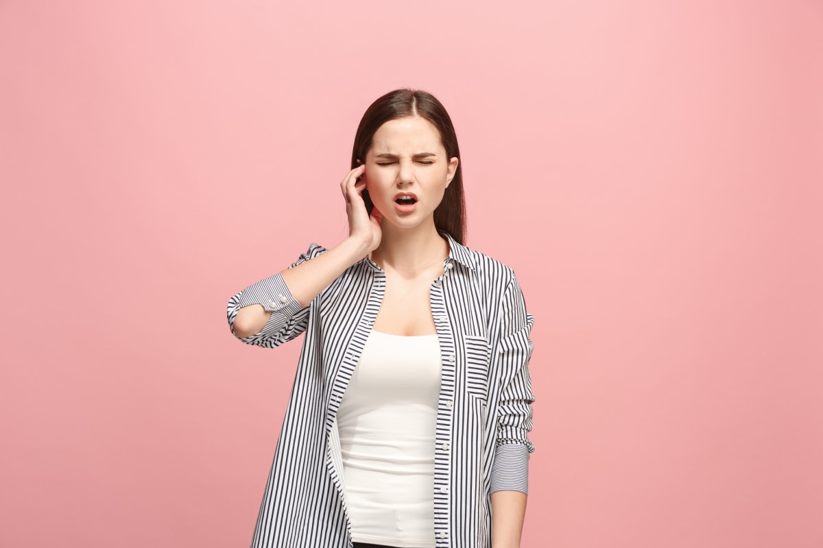 A una mujer le duele la oreja y se la está sujetando. Está de pie. Fondo rosa.