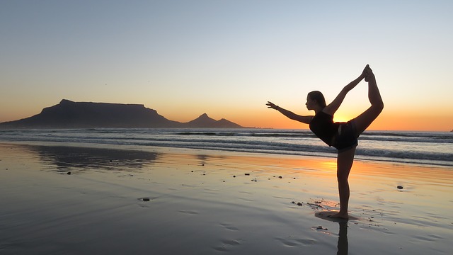 Una mujer practica yoga en la playa. El sol se ha puesto, aún hay luz. Está de pie sobre una pierna. Es marea baja. La arena está bajo una fina capa de agua.