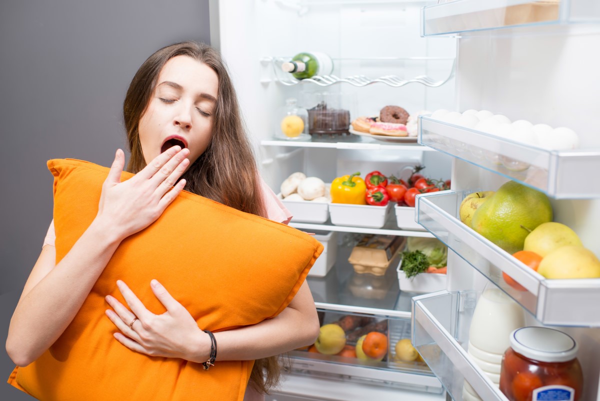 Una mujer durmiendo junto a un frigorífico, lo que ilustra los hábitos alimentarios como factor que influye en la calidad del sueño.