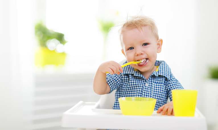 Un niño pequeño sentado en una sillita de bebé, dándose de comer. Cuchara, taza y cuenco amarillos.